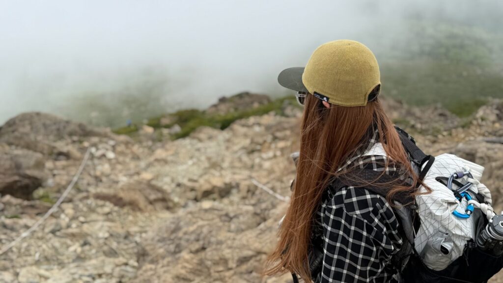 U.L. Merino X Capを被ってガスのかかる岩場の登山道を見下ろす後ろ姿